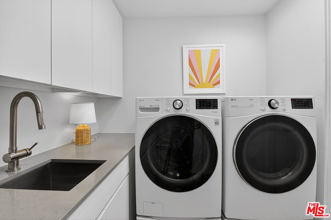 This clean and modern laundry room features a side-by-side washer and dryer set with dark circular doors. To the left, a sleek sink is integrated into a stone countertop, complemented by white cabinetry and a minimalist faucet. A small, textured lamp and a vibrant, sun-themed piece of wall art add a touch of personality to the bright, functional space.