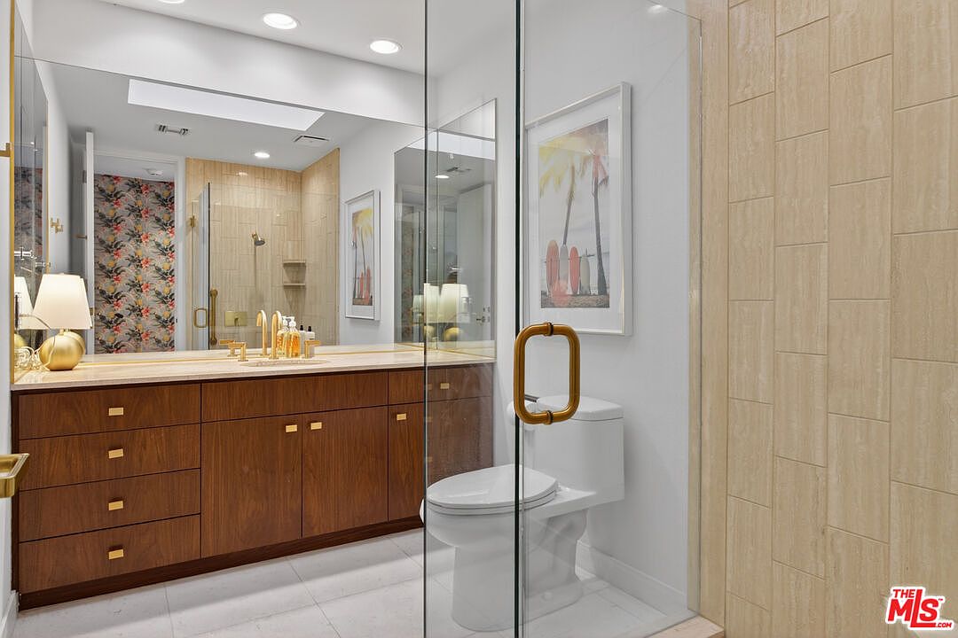This modern bathroom features a wood-toned vanity with a light-colored countertop and gold-toned fixtures. A glass-enclosed shower area is positioned next to a white toilet, while a large mirror reflects the space and a framed piece of beach-themed artwork. The room is accented with patterned wallpaper in the background and light-colored wall tiles, creating a bright and contemporary aesthetic.
