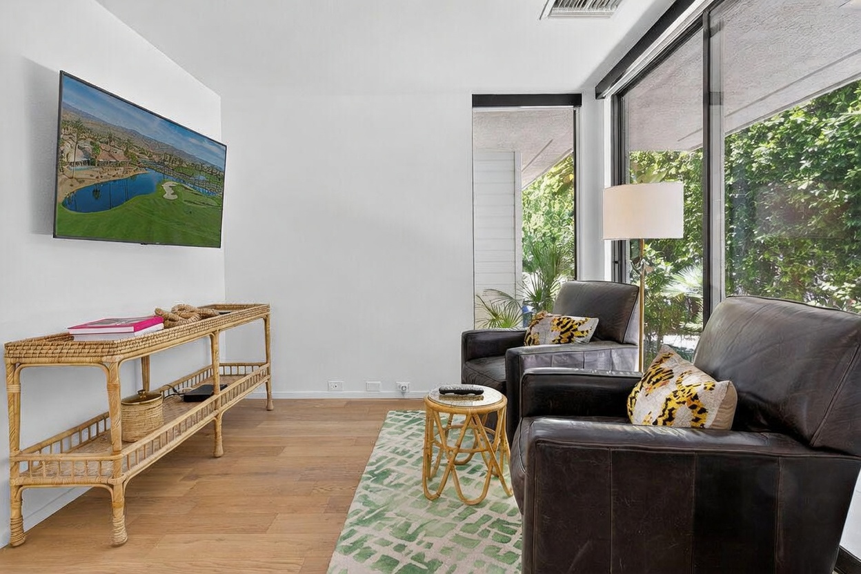This bright living area features light wood flooring and a wall-mounted television above a long, woven rattan console table. Two dark leather armchairs are positioned near large windows that provide a view of lush greenery outside. A small, matching rattan side table and a patterned area rug complete the cozy, relaxed seating arrangement.