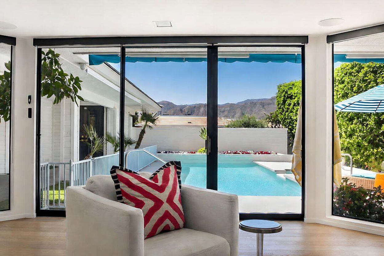 This interior view captures a bright living space looking out through expansive glass sliding doors toward a private backyard pool. A comfortable armchair with a bold red and white patterned pillow sits in the foreground, while the background reveals a clear blue pool, a white privacy wall, and distant desert mountains. The scene emphasizes a seamless indoor-outdoor connection, highlighting the home's resort-like atmosphere and natural light.