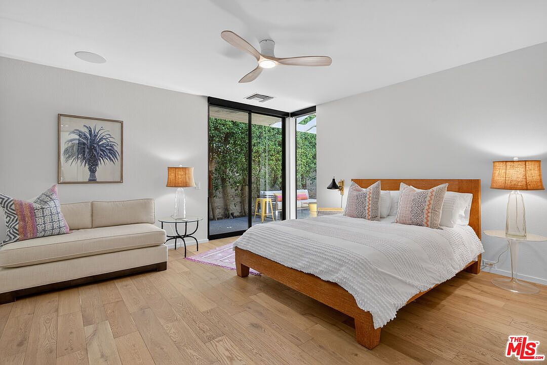 This bright and airy bedroom features light wood flooring and a neutral color palette, centered around a wooden bed frame with a white textured duvet. To the left, a beige sofa provides a cozy seating area, while large sliding glass doors offer a seamless transition to an outdoor patio space. The room is illuminated by a modern ceiling fan and matching bedside lamps, creating a serene and inviting atmosphere.
