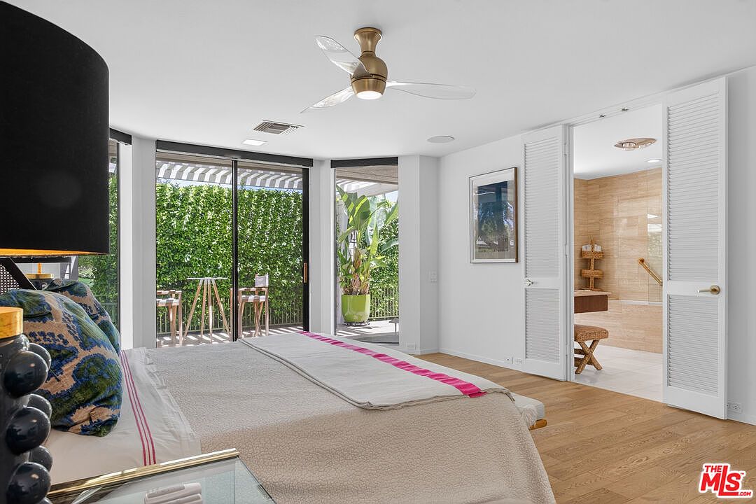 This bright primary bedroom features light wood flooring and a neutral color palette, centered around a bed with patterned pillows and a pink-accented throw. Large sliding glass doors provide access to an outdoor patio area, while white louvered doors open into an adjacent bathroom. A modern ceiling fan hangs above, and the room is accented by a framed piece of art on the wall.