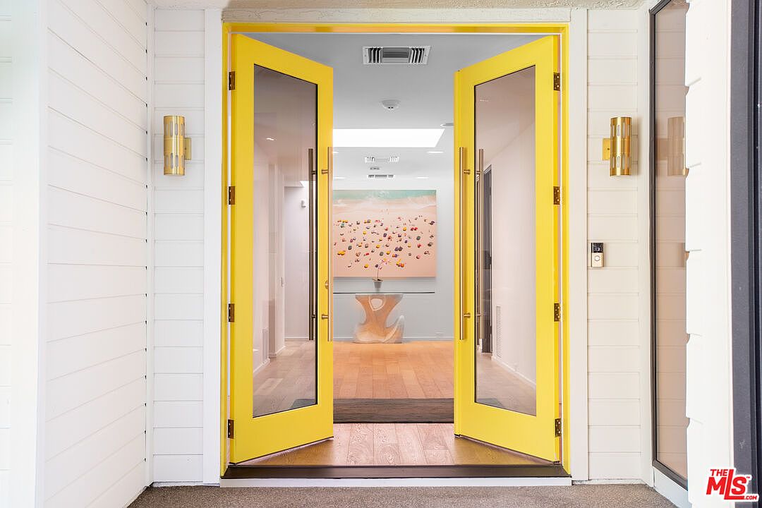 This striking entryway features a pair of vibrant yellow double doors with large glass panels, creating a bold and welcoming focal point. The doors are flanked by modern, vertical gold-toned wall sconces and white horizontal siding, leading into a bright, open foyer. Through the doorway, a glimpse of a light-filled interior reveals a contemporary art piece and a sculptural console table, establishing a sophisticated and artistic aesthetic.