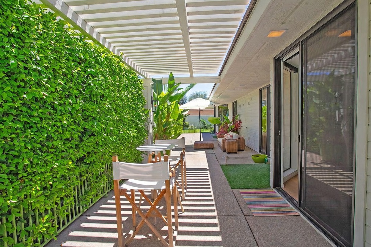 This inviting outdoor patio features a white pergola overhead that casts rhythmic shadows across the concrete floor. The space is furnished with a small bistro set and a comfortable seating area further down, all bordered by a lush, tall green hedge and tropical plants. Large sliding glass doors provide seamless access from the interior to this serene, sun-drenched living space.