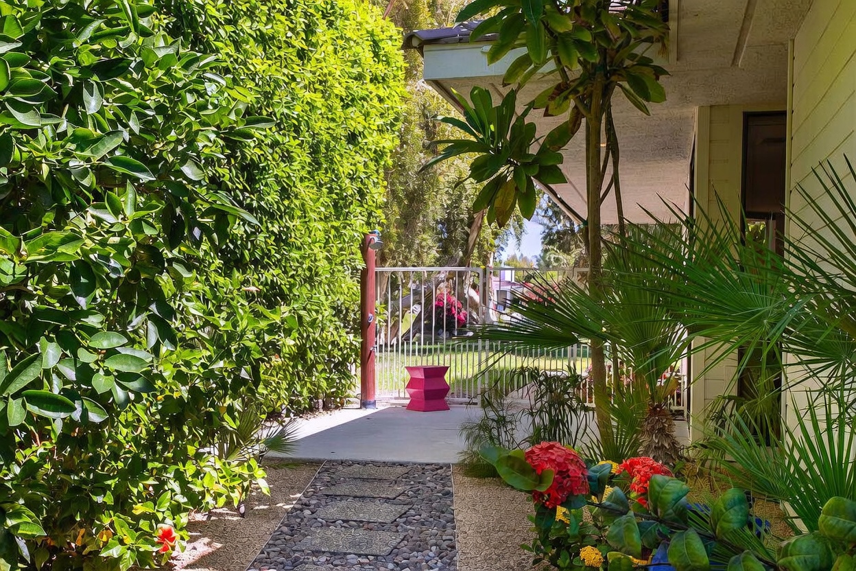 This outdoor space features a concrete patio area shaded by a roof overhang, leading to a stone-paved walkway through a lush garden. A tall, slender outdoor shower stands near a white metal fence, while vibrant red flowers and tropical greenery frame the scene. The perspective is from the edge of the patio, looking out toward the garden and the fence, creating a serene and private backyard atmosphere.