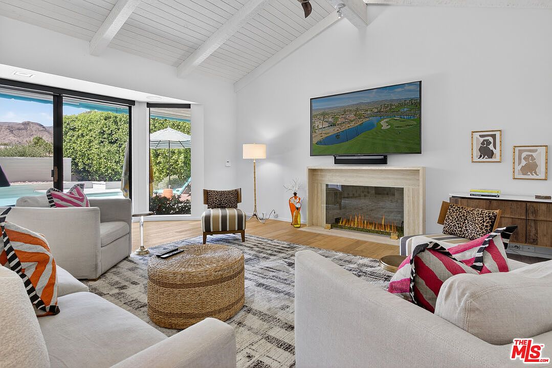 This bright and airy living room features a vaulted ceiling with exposed beams and a large, light-toned stone fireplace surround housing a linear fire feature. The space is furnished with comfortable, neutral-toned seating, a textured round ottoman, and a patterned area rug, all centered around the television mounted above the fireplace. Large sliding glass doors provide a seamless connection to the outdoor pool area, filling the room with natural light and offering a scenic view of the landscape.