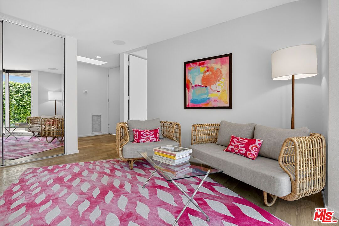This bright living area features a contemporary aesthetic with a bold, pink-and-white patterned area rug as the focal point. The space is furnished with a light-toned sofa and matching armchair, both accented with vibrant patterned throw pillows and featuring woven rattan frames. A glass-topped coffee table sits in the center, while a tall floor lamp and colorful abstract wall art add warmth and personality to the room.