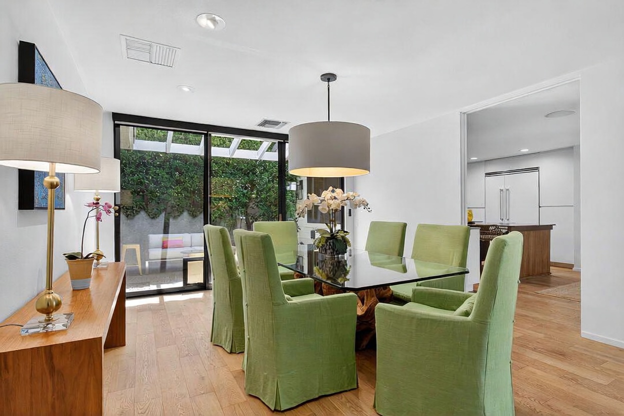 This bright dining room features a glass-topped table surrounded by six lime-green upholstered chairs, centered beneath a large, modern drum pendant light. A long wooden console table sits against the left wall, topped with a lamp and a potted plant, while a sliding glass door provides a view of the outdoor patio. The space is finished with light wood flooring and white walls, creating an airy and inviting atmosphere that flows into the adjacent kitchen.