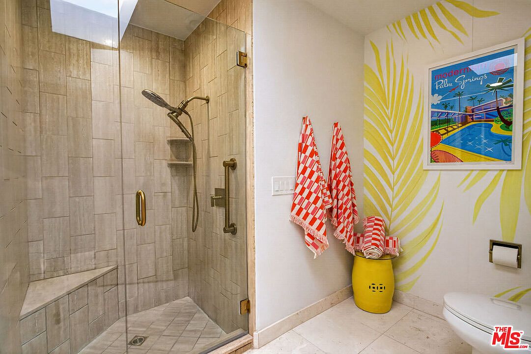 This vibrant bathroom features a walk-in shower with a glass enclosure, tiled walls, and a built-in bench. The room is accented by a playful palm-leaf patterned wallpaper, a bright yellow stool, and red-and-white checkered towels. The perspective captures the shower area alongside a decorative wall, creating a cheerful and modern aesthetic.