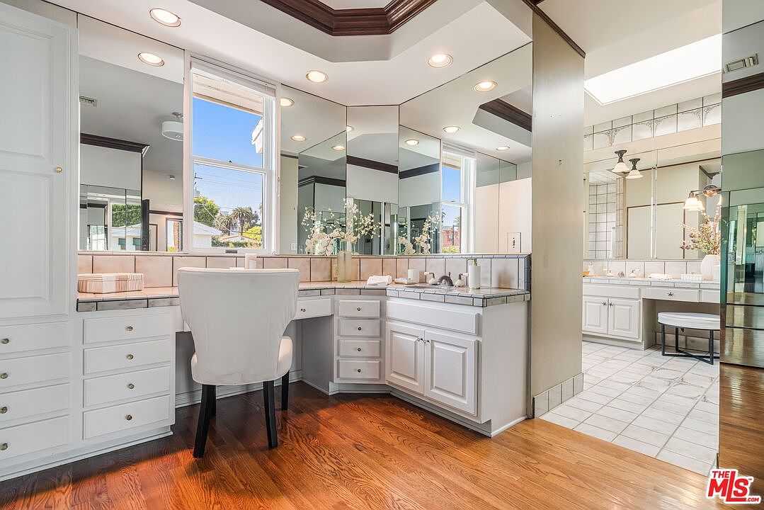 This primary bathroom features a spacious vanity area with white cabinetry, tiled countertops, and a large, multi-paneled mirror that reflects the room's bright, airy atmosphere. A white upholstered chair is tucked under the vanity, and the space transitions into a secondary tiled bathroom area visible through an open doorway. The room is finished with warm-toned wood flooring and recessed lighting, creating a classic and functional aesthetic.