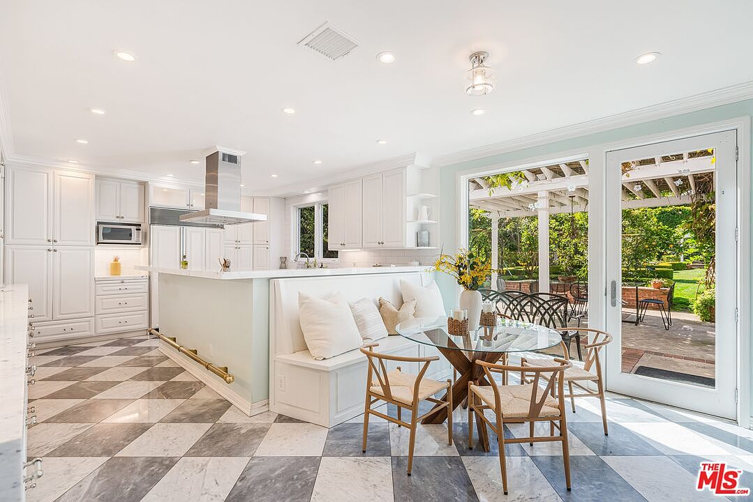 This bright and airy kitchen features a large central island with a breakfast nook bench, paired with a glass-topped dining table and light wood chairs. The space is defined by white cabinetry, a stainless steel range hood, and a distinctive checkered stone floor. Large glass doors open directly to a lush, sunlit garden patio, creating a seamless transition between indoor and outdoor living areas.