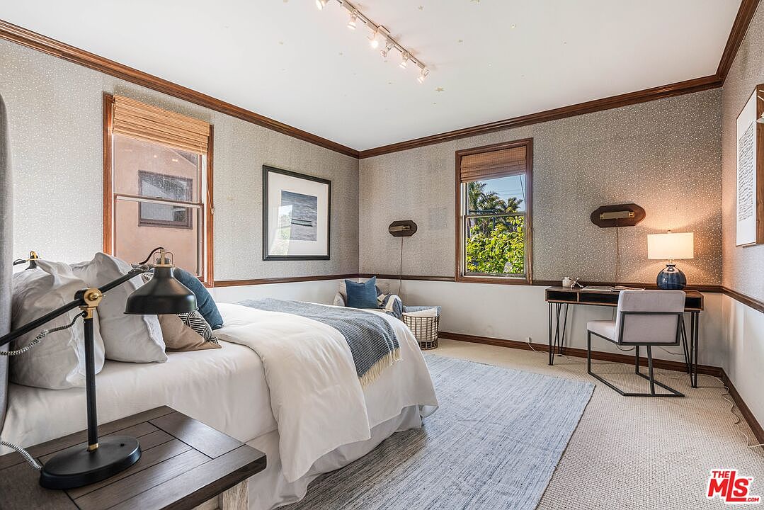 This well-appointed bedroom features a neutral color palette with patterned wallpaper and dark wood trim. The room is furnished with a bed dressed in white linens, a desk with a modern chair, and a small seating area by the window, creating a functional and inviting space. The perspective is from the doorway, capturing the entire room layout including the track lighting and the natural light streaming through the windows.