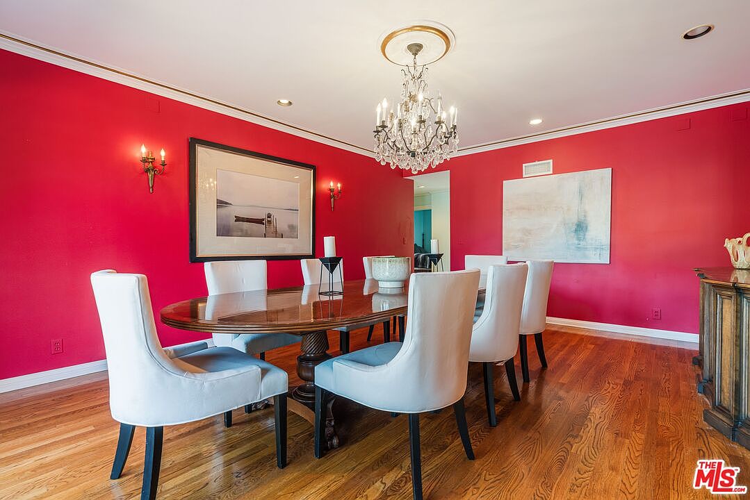 This formal dining room features bold red walls contrasted by white-upholstered chairs surrounding a large, dark wood oval table. A crystal chandelier hangs from a decorative ceiling medallion, serving as the focal point above the table, while two pieces of framed artwork adorn the walls. The space is finished with warm-toned wood flooring and crown molding, creating a sophisticated and traditional atmosphere.