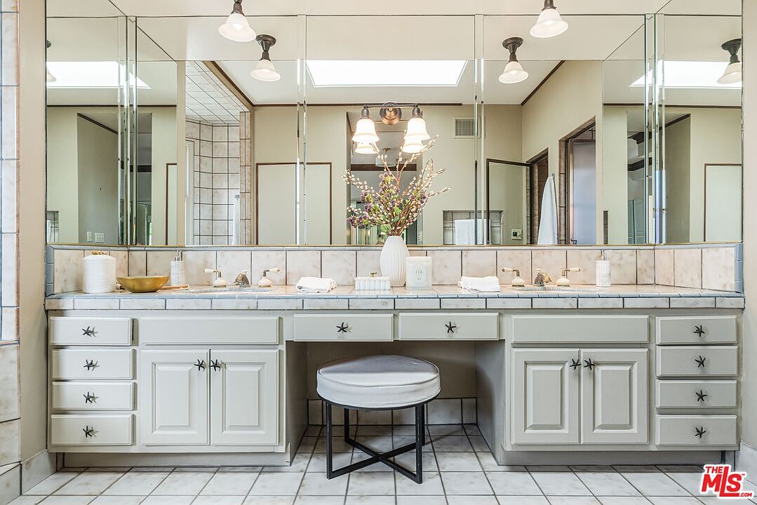 This primary bathroom features a double vanity with light-colored cabinetry, a central makeup vanity area with a stool, and a large mirror spanning the wall. The space is illuminated by multiple light fixtures and includes a tiled countertop and backsplash. The perspective is a straight-on, eye-level shot capturing the symmetry and functional layout of the vanity area.