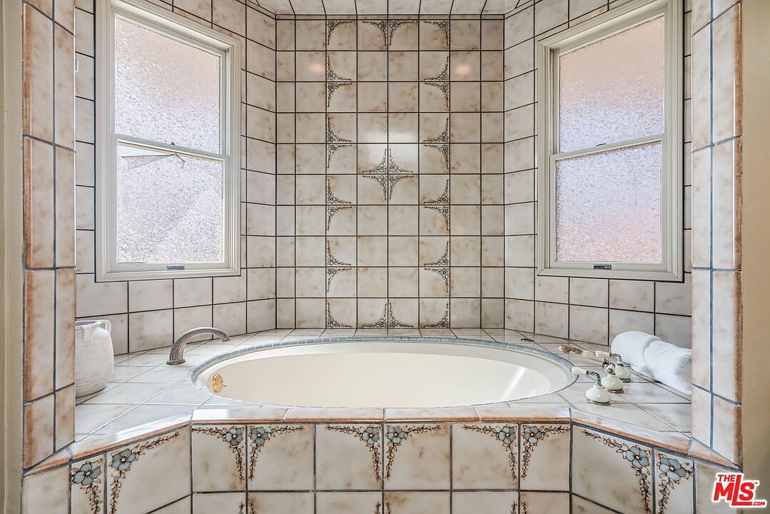 This primary bathroom features a large, circular soaking tub set into a tiled deck, positioned beneath two windows that provide natural light. The walls are adorned with patterned ceramic tiles, including decorative floral accents and a central star motif. The space is framed by a tiled surround, creating a classic, spa-like atmosphere.