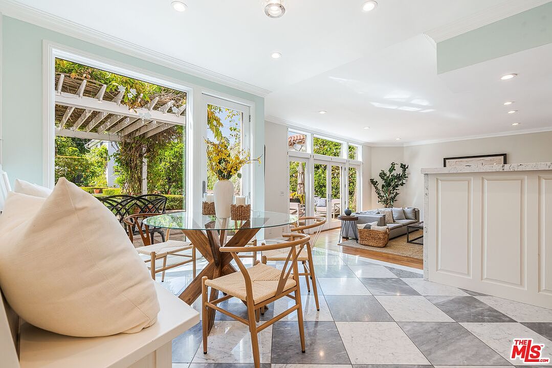 This bright and airy dining area features a glass-topped table surrounded by light wood chairs, set against a backdrop of large windows that look out onto a lush garden. The space is finished with a classic checkerboard tile floor and flows seamlessly into an adjacent living area, creating an open-concept feel. The neutral color palette and abundant natural light contribute to a sophisticated and inviting atmosphere.
