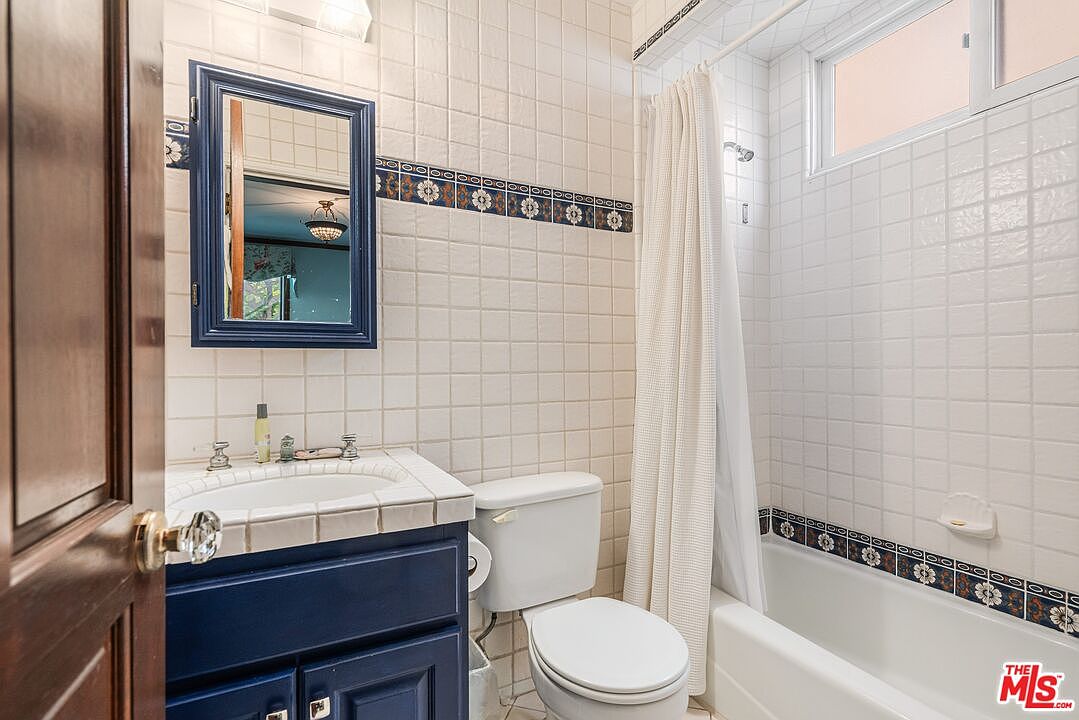 This compact bathroom features a navy blue vanity with a tiled countertop and a matching framed mirror. The space is finished with light-colored wall tiles accented by a decorative floral border, and includes a standard toilet and a shower-tub combination with a white curtain. The perspective is a straight-on view from the doorway, highlighting the functional layout of the room.