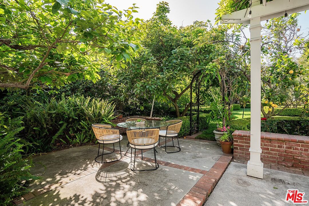 This charming outdoor patio space features a glass-topped dining table surrounded by four modern, woven-style chairs. The area is nestled under the lush canopy of mature trees and greenery, creating a private and serene garden atmosphere. A white pergola post stands to the right, adjacent to a low brick wall, providing a classic and inviting setting for al fresco dining.