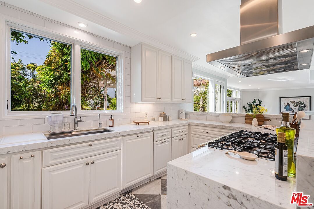 This bright and airy kitchen features white cabinetry paired with light-toned stone countertops and a classic subway tile backsplash. A large island with a gas cooktop and a stainless steel range hood serves as the centerpiece, while a window above the sink provides a view of the lush greenery outside. The space is finished with patterned tile flooring, creating a clean and inviting atmosphere suitable for a modern home.