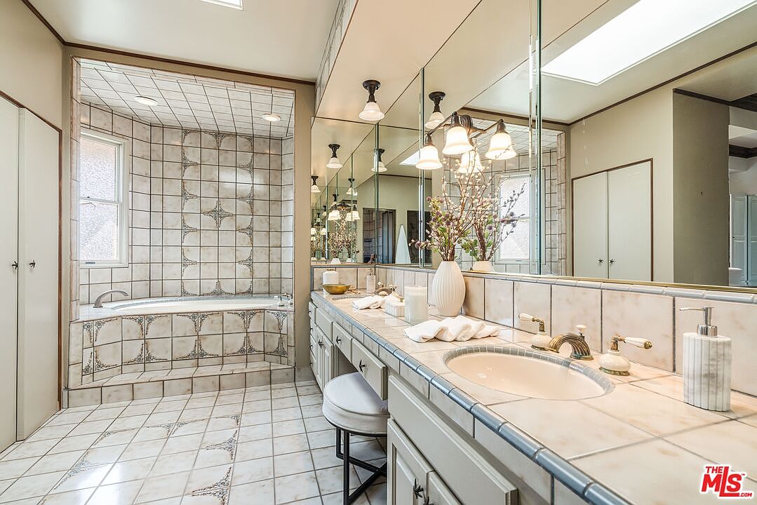 This spacious primary bathroom features a large soaking tub set within a tiled alcove and a long vanity with dual sinks. The room is finished with light-toned patterned tile flooring and walls, complemented by an expansive wall-to-wall mirror that enhances the sense of space. A small stool sits tucked under the vanity, and the area is illuminated by a combination of recessed lighting and a decorative multi-light fixture.