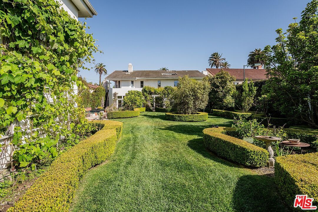 This expansive, manicured backyard features a lush green lawn framed by neatly trimmed boxwood hedges and mature landscaping. A white two-story residence serves as the backdrop, with climbing vines adorning the exterior walls and a central tree providing a focal point in the garden. The perspective is a wide-angle ground-level shot looking toward the house, capturing the serene and well-maintained outdoor living space.