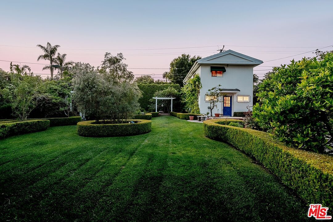 This image captures a serene, manicured backyard featuring a lush green lawn framed by neatly trimmed boxwood hedges. A small, two-story white structure with a vibrant blue door serves as a focal point, complemented by a nearby pergola and mature trees. The perspective is a wide, eye-level shot that emphasizes the peaceful, park-like atmosphere of the property.