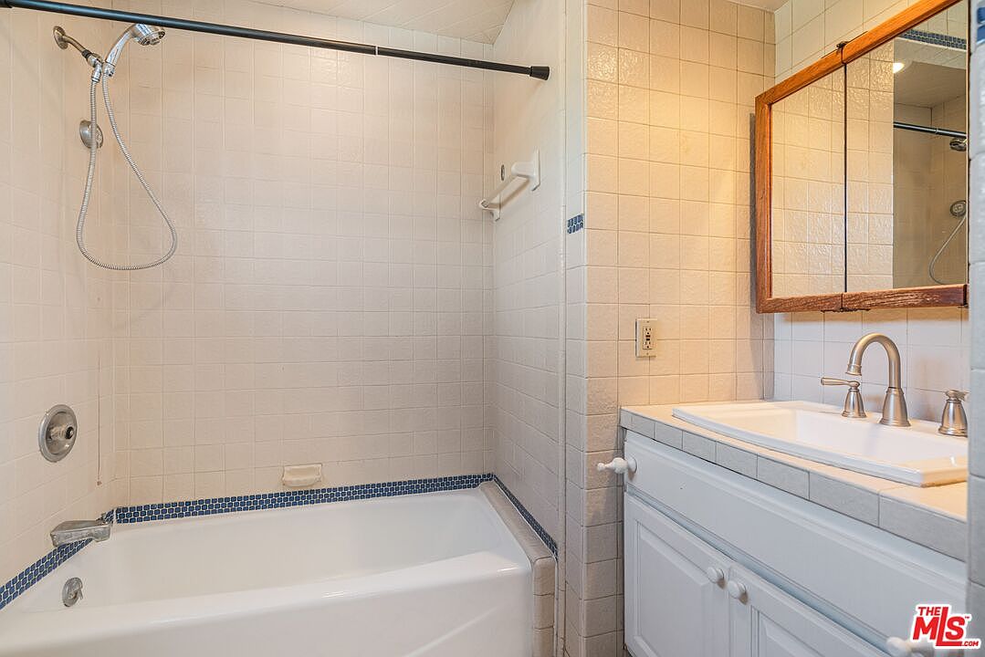 This bathroom features a combination tub and shower unit with a handheld showerhead and a decorative blue-tiled accent border. A white vanity with a rectangular sink and a wooden medicine cabinet mirror provides functional storage. The space is finished with light-colored wall tiles and a neutral countertop, offering a clean and practical layout.