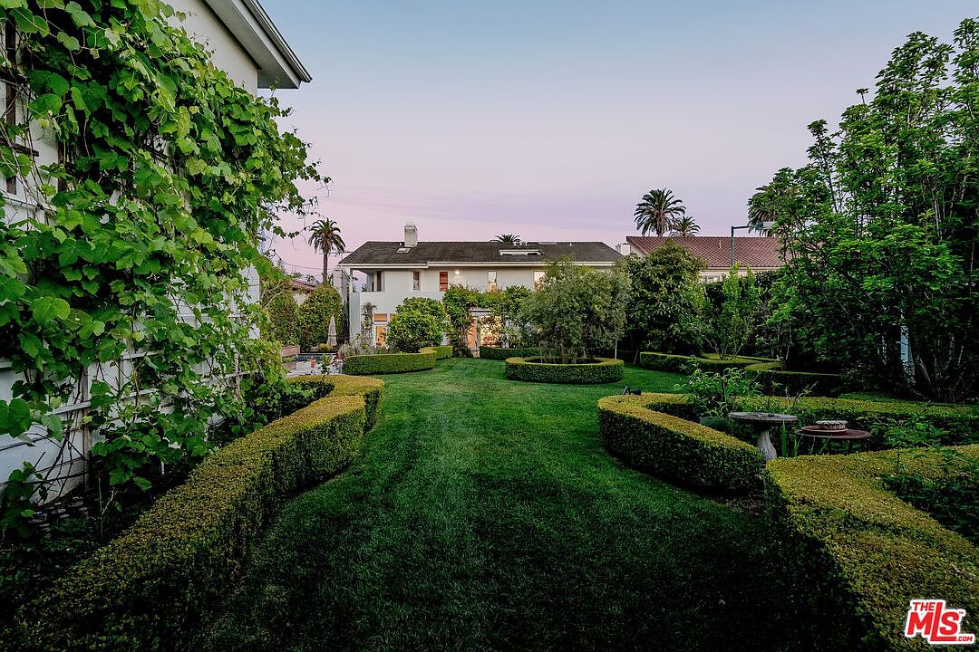 This expansive backyard features a meticulously manicured lawn framed by structured, low-profile boxwood hedges. A lush climbing vine covers the side of the house on the left, while mature trees and shrubs provide a sense of privacy and depth to the landscape. The perspective is a ground-level view looking toward the rear facade of the two-story home during the soft light of dusk.