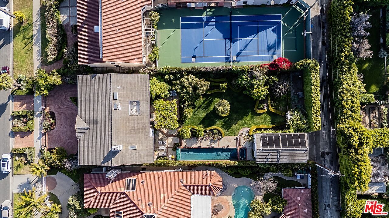 This high-angle aerial view captures a residential property featuring a private tennis court, a lush green lawn with manicured landscaping, and a rectangular swimming pool. The layout showcases a multi-level home with a tiled roof, surrounded by dense perimeter hedges that provide significant privacy. The perspective highlights the expansive outdoor living space and the integration of recreational amenities within the property grounds.