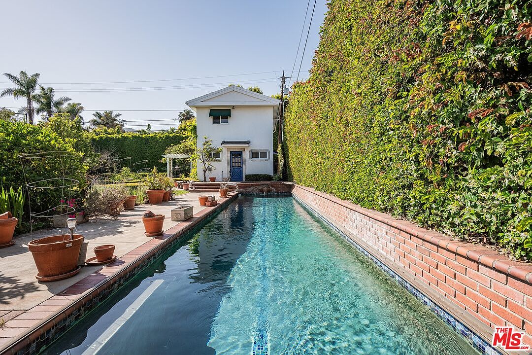 This image captures a long, narrow lap pool stretching toward a charming white two-story structure at the end of the yard. A brick-lined walkway runs alongside the pool, adorned with numerous potted plants and a tall, lush green hedge that provides significant privacy. The perspective is from the edge of the pool, looking down the length of the water toward the building, creating a serene and private outdoor retreat.