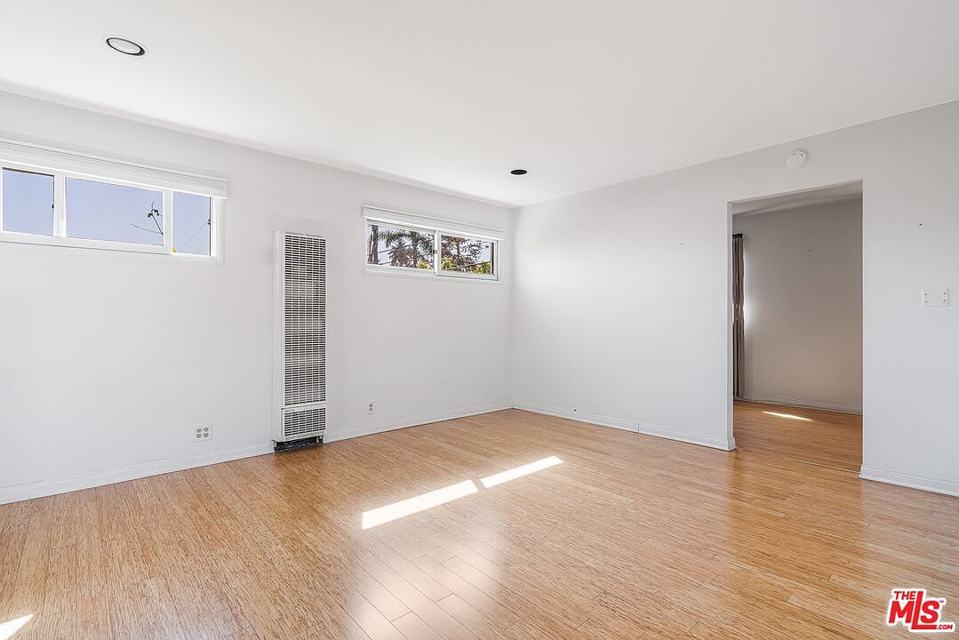 This bright, minimalist living room features light-toned wood flooring and crisp white walls, creating an airy and open atmosphere. The space is illuminated by natural light streaming through two windows, with a wall-mounted heater unit positioned between them. A doorway on the right leads into an adjacent room, providing a sense of flow and connectivity throughout the home.