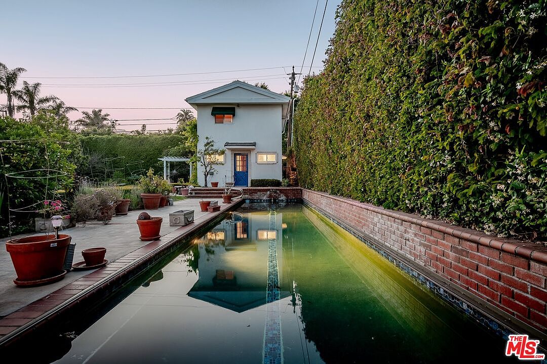 This outdoor space features a long, narrow lap pool bordered by a brick wall and a paved patio area filled with potted plants. A detached, two-story white structure with a vibrant blue door stands at the end of the pool, framed by a tall, dense hedge that provides significant privacy. The perspective is a low-angle shot looking down the length of the pool, emphasizing the elongated layout and the serene, garden-like atmosphere.
