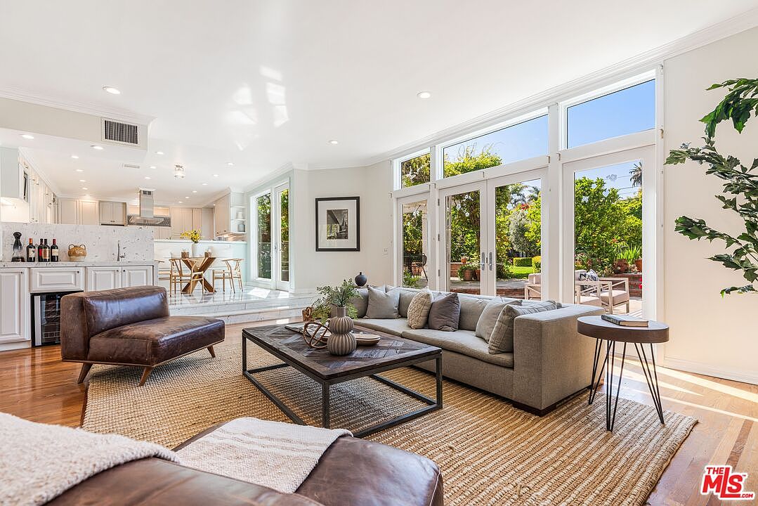 This bright and airy living room features a comfortable gray sofa and a leather armchair arranged around a dark wood coffee table on a large textured rug. The space is bathed in natural light from expansive glass doors that open to a lush backyard, creating a seamless indoor-outdoor flow. In the background, a raised dining area and a modern kitchen with white cabinetry are visible, highlighting an open-concept floor plan.
