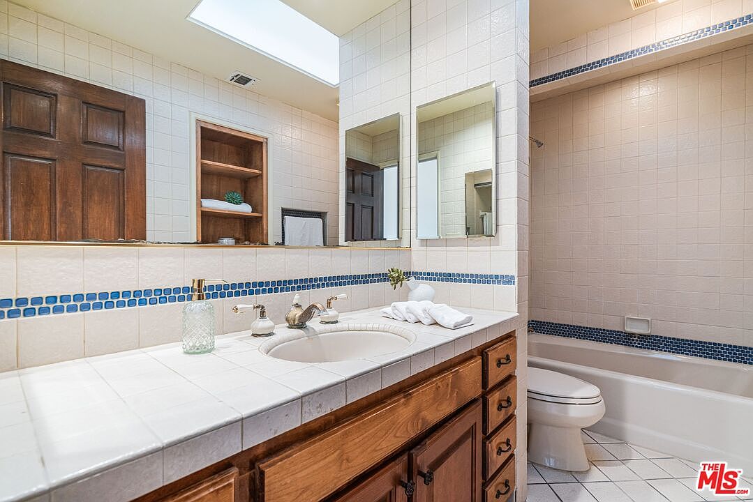 This bathroom features a vanity with a light-toned wood cabinet, a white tiled countertop, and a single sink. A large mirror spans the wall above the vanity, reflecting a built-in shelving unit and a doorway, while a tiled shower-tub combination is positioned to the right. The space is accented with a decorative blue and white patterned tile border along the wall, and a skylight above provides natural illumination.