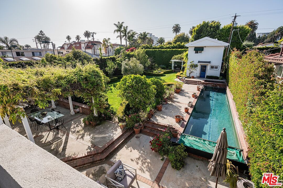 This elevated perspective captures a lush, private backyard oasis featuring a long, rectangular swimming pool bordered by a concrete patio and mature landscaping. To the left, a shaded pergola covers an outdoor dining area, while a detached white structure with a blue door sits at the far end of the garden. The scene is framed by tall, manicured hedges and trees, creating a serene and secluded atmosphere perfect for outdoor entertaining.