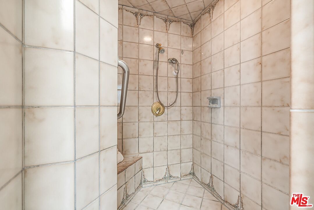 This shower enclosure features light-toned, square-patterned wall tiles with decorative floral accents near the corners. The space includes a built-in tiled bench, a handheld shower fixture, and a metal grab bar for accessibility. The perspective is from the entrance of the shower, looking toward the back wall and the integrated seating area.