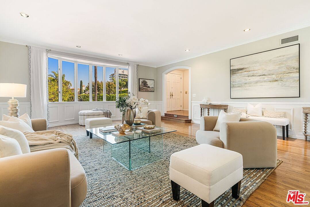 This bright and airy living room features a neutral color palette with plush cream-colored sofas and a central glass-topped coffee table. A large window arrangement allows for abundant natural light, while a textured area rug anchors the seating area, which is complemented by a large piece of abstract wall art. The space flows seamlessly into an adjacent hallway, creating an open and inviting atmosphere suitable for formal entertaining.