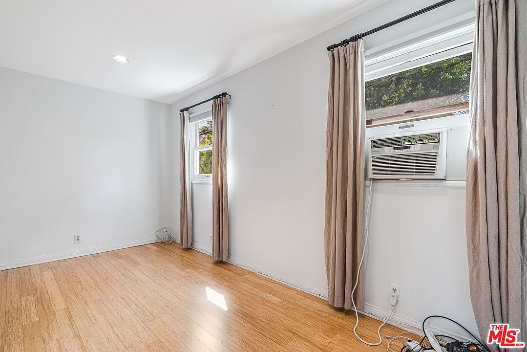 This bright, minimalist bedroom features light-colored walls and warm-toned wood flooring. The room is illuminated by recessed lighting and natural light streaming through two windows dressed with simple, neutral-toned curtains. A window-mounted air conditioning unit is visible, and the space offers a clean, blank canvas ready for personalization.