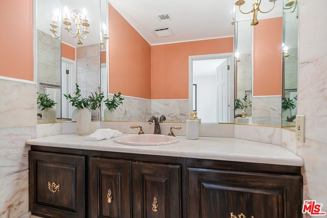 This bathroom features a vanity with a dark wood cabinet and a light-colored stone countertop, accented by a pastel pink sink basin. The walls are finished with a combination of peach-toned paint and light-colored stone wainscoting, complemented by large mirrors that reflect the space. The perspective is a straight-on, eye-level shot capturing the vanity area, highlighting the traditional design elements and decorative lighting fixtures.