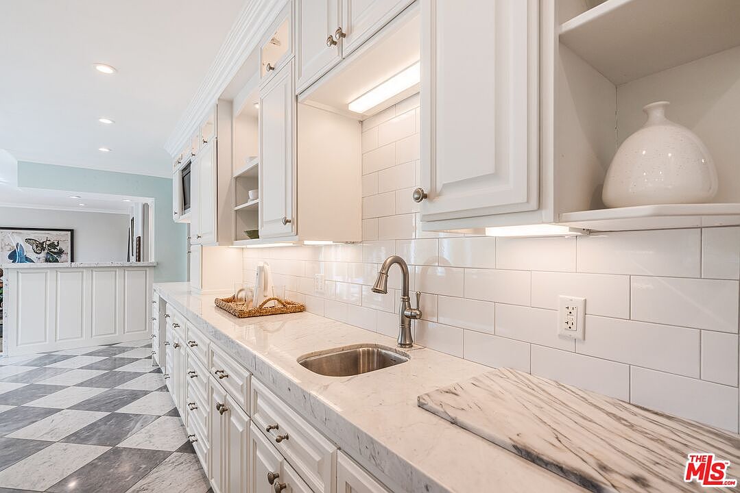 This bright kitchen features white cabinetry, a light-toned stone countertop, and a classic white subway tile backsplash. A stainless steel sink with a gooseneck faucet is centered on the counter, while a marble-patterned cutting board rests on the surface. The space is finished with a distinctive black and white checkered floor, creating a clean and elegant aesthetic.