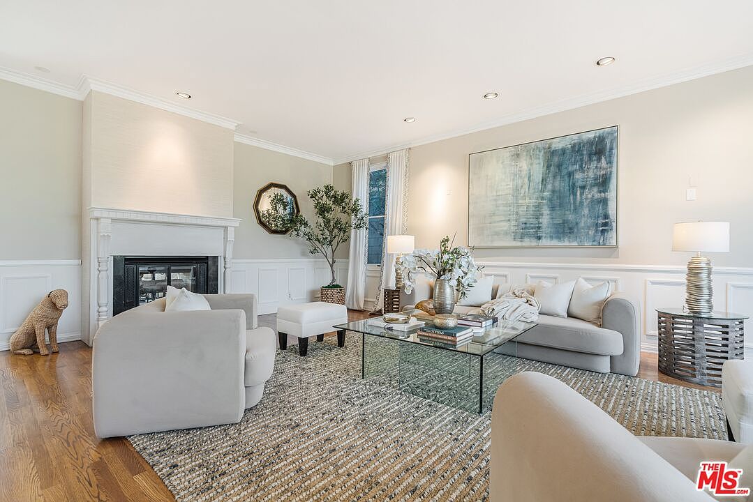 This bright and airy living room features a neutral color palette with light-toned walls and wainscoting. The space is anchored by a central fireplace with a classic mantel, a glass-topped coffee table, and comfortable seating arranged on a textured area rug. A large abstract painting hangs above the sofa, while a decorative dog statue and a potted tree add character to the room.
