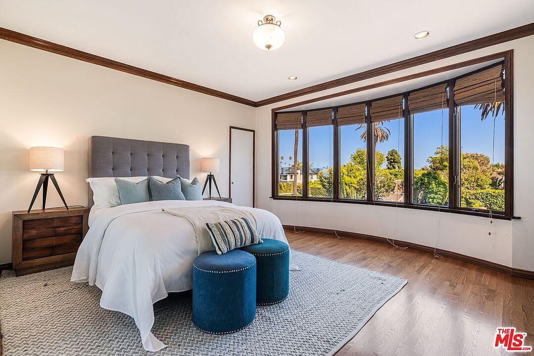 This bright and airy primary bedroom features a central bed with a grey tufted headboard, flanked by two wooden nightstands with modern lamps. A large bay window with woven shades provides ample natural light and views of the surrounding greenery, while two blue upholstered ottomans sit at the foot of the bed. The room is finished with warm wood flooring and a textured area rug, creating a comfortable and inviting atmosphere.