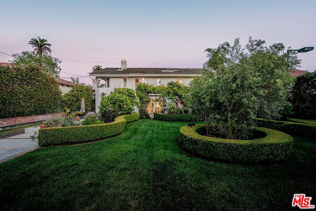 This rear view of a two-story residence showcases a well-manicured lawn framed by neatly trimmed boxwood hedges and mature trees. The home features a light-colored exterior with a central entrance area adorned with climbing vines, creating a serene and private backyard atmosphere. The perspective is captured from the lawn, emphasizing the lush landscaping and the peaceful transition between the garden and the house.