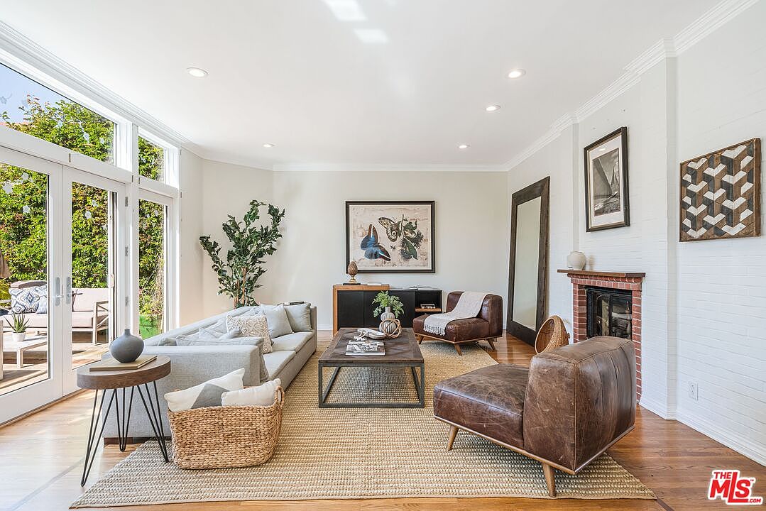 This bright and airy living room features a neutral color palette, hardwood flooring, and a large area rug that anchors the seating arrangement. The space includes a light-colored sofa, two leather armchairs, and a wooden coffee table, all centered around a brick-surround fireplace with a wooden mantel. Large glass doors provide ample natural light and a view of the outdoor patio, creating a seamless transition between indoor and outdoor living.