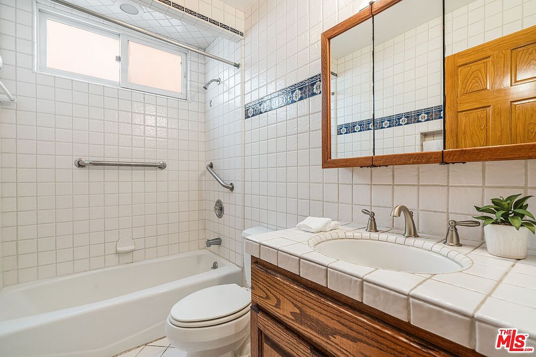 This bathroom features a classic design with white square wall tiles accented by a decorative blue and white patterned border. The space includes a combination bathtub and shower with a sliding window, a wooden vanity with a tiled countertop, and a medicine cabinet with mirrored doors. A small potted plant adds a touch of greenery to the vanity, creating a functional and traditional aesthetic.