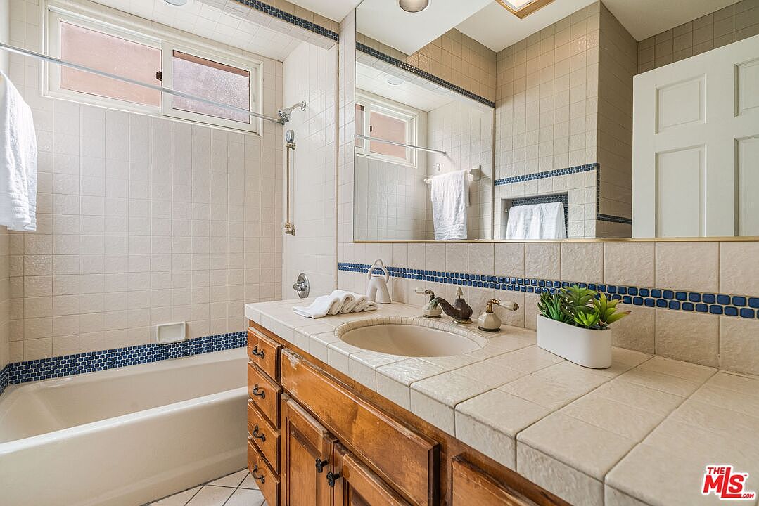 This bathroom features a vanity with a tiled countertop and light wood cabinetry, paired with a large wall-mounted mirror that enhances the sense of space. A combination bathtub and shower area is enclosed by a tiled wall with a decorative blue accent border. The room is well-lit by natural light from a window above the tub and overhead lighting, creating a functional and traditional aesthetic.