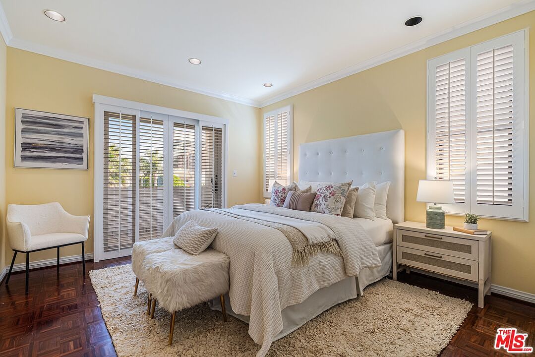 This bright and airy primary bedroom features soft yellow walls, a large white tufted headboard, and a plush area rug over dark wood flooring. A set of sliding glass doors with plantation shutters allows for natural light, while a matching nightstand and a textured bench at the foot of the bed complete the space. The room offers a serene and inviting atmosphere with a neutral color palette.