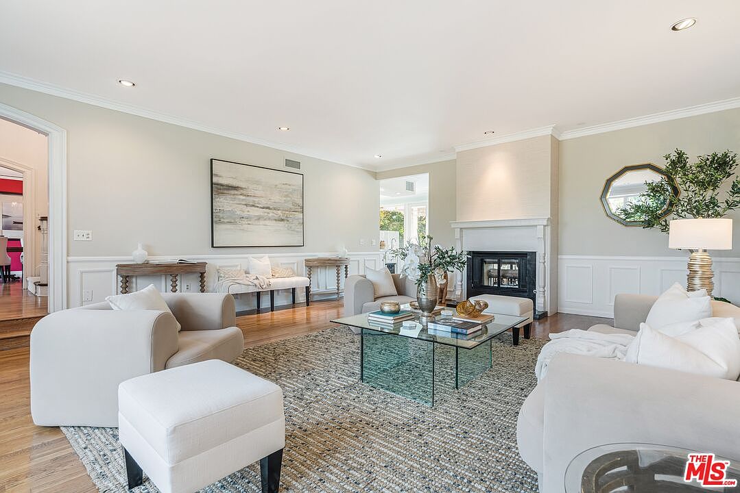 This bright and airy living room features a neutral color palette with comfortable seating arranged around a glass-topped coffee table. A prominent fireplace with a white mantel serves as a focal point, complemented by a large abstract painting on the adjacent wall and a decorative mirror above a lamp. The space is finished with light wood flooring and a textured area rug, creating a sophisticated and inviting atmosphere.