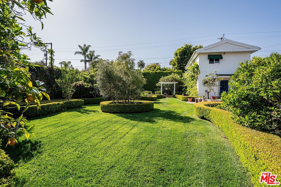 This expansive, manicured backyard features a lush green lawn framed by neatly trimmed hedges and mature trees. A white pergola stands in the distance, providing a focal point at the end of the garden path, while a white two-story structure with a blue door sits to the right. The scene is captured from a ground-level perspective, emphasizing the serene and well-maintained outdoor living space.