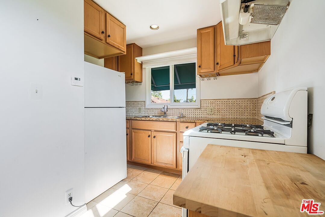 This kitchen features light wood cabinetry, a white refrigerator, and a gas range, creating a functional and compact workspace. A window above the sink provides natural light, while a wooden butcher-block style counter extension adds extra prep space. The room is finished with light-colored tile flooring and a neutral backsplash, offering a clean and traditional aesthetic.